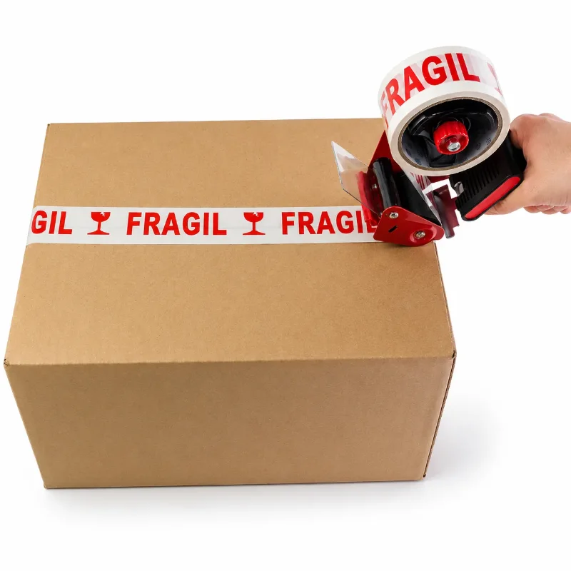 Una mano con portacintas rojo y negro pega cinta blanca con la palabra FRAGIL sobre la unión de las solapas de una caja